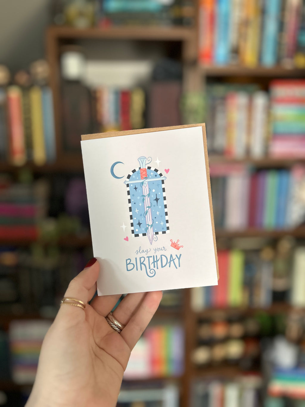 Fantasy-inspired birthday greeting card featuring a magical sword surrounded by stars, hearts, a crescent moon, and the message "Slay Your Birthday" on a soft neutral background.