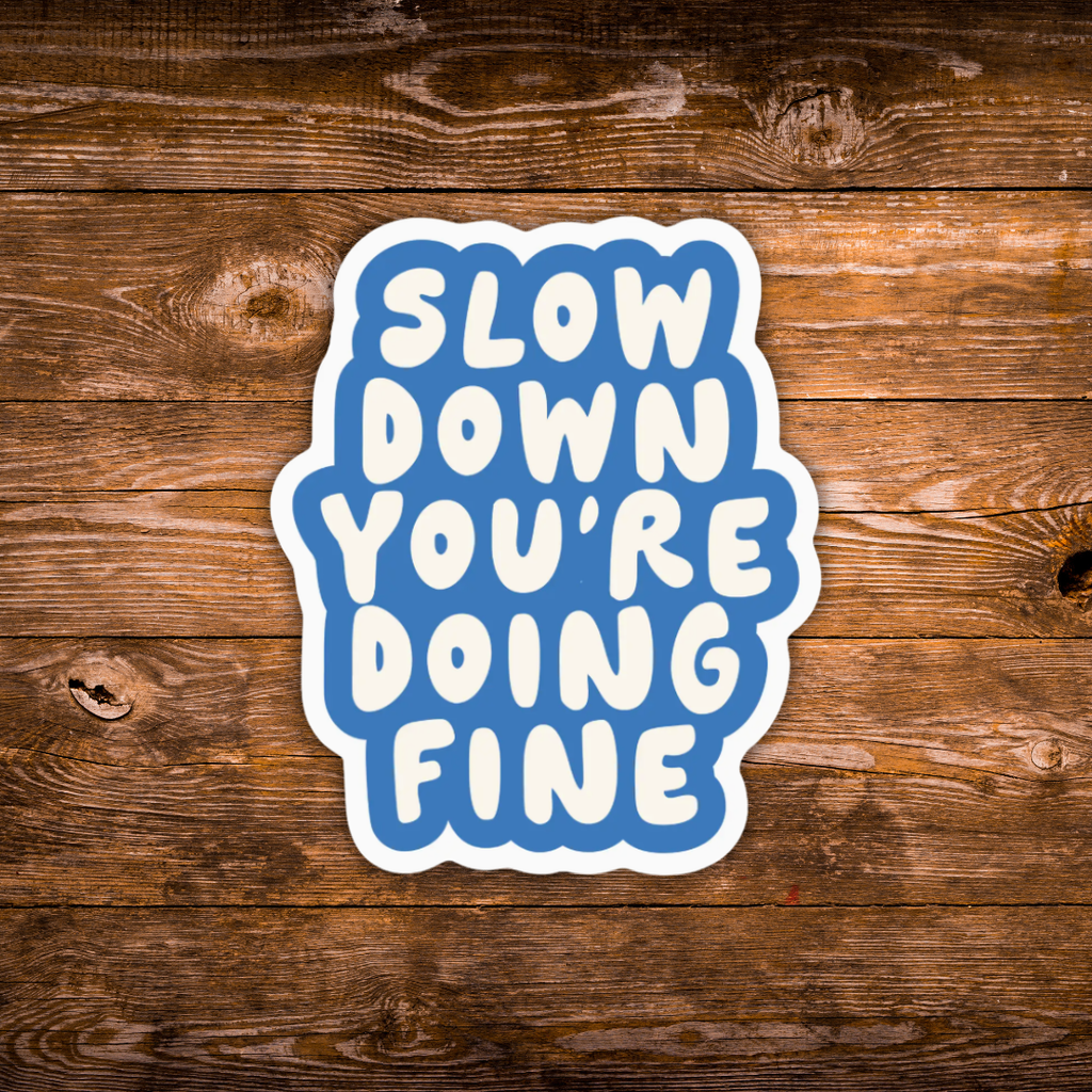 A die-cut sticker with the text 'SLOW DOWN YOU'RE DOING FINE' in white rounded letters on a blue background with a white border, displayed on a rustic brown wooden surface.
