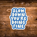 A die-cut sticker with the text 'SLOW DOWN YOU'RE DOING FINE' in white rounded letters on a blue background with a white border, displayed on a rustic brown wooden surface.