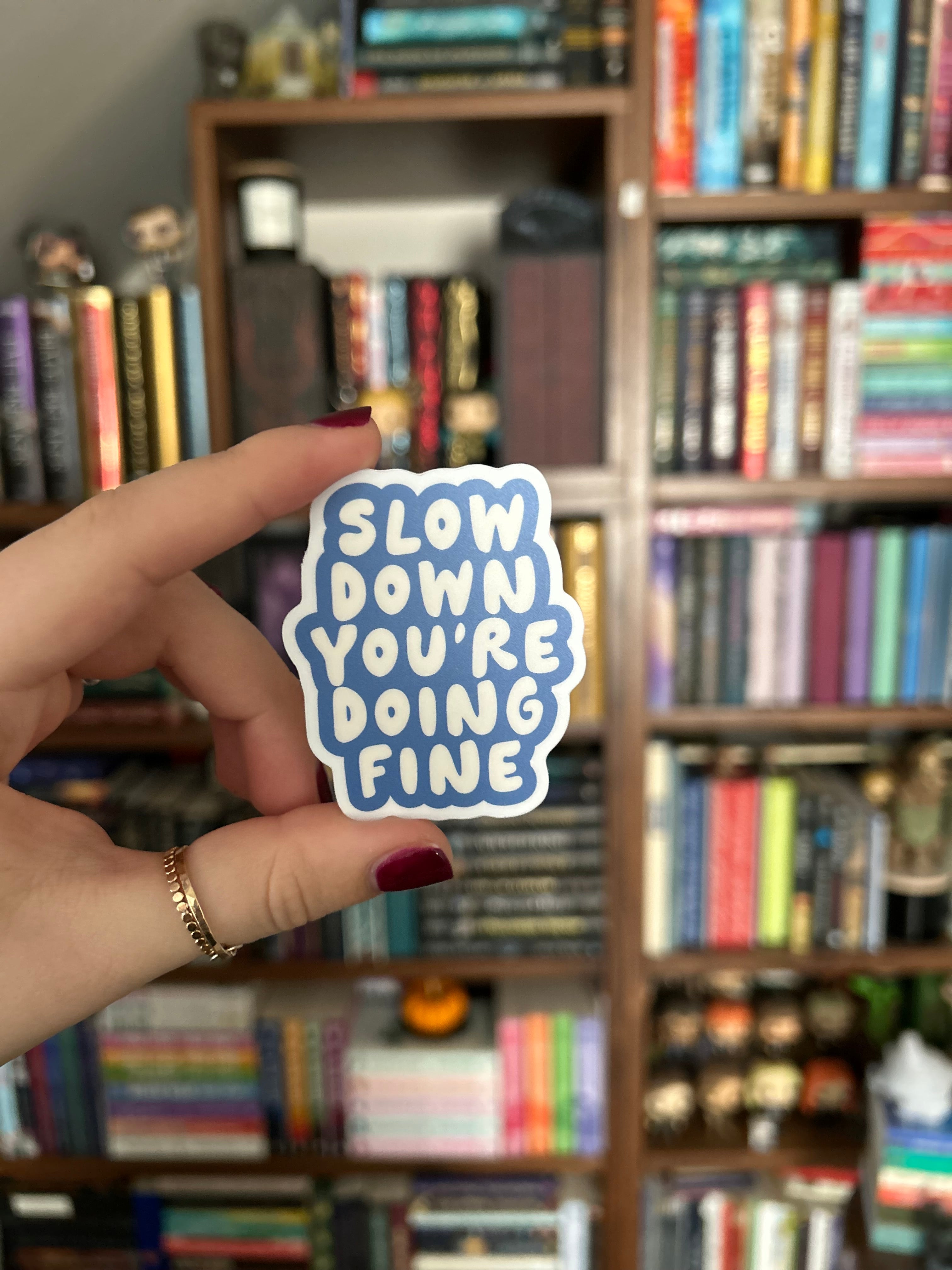 A hand holding a die-cut sticker with the text 'SLOW DOWN YOU'RE DOING FINE' in white letters on a blue cloud-like outline, set against a blurred background of bookshelves.