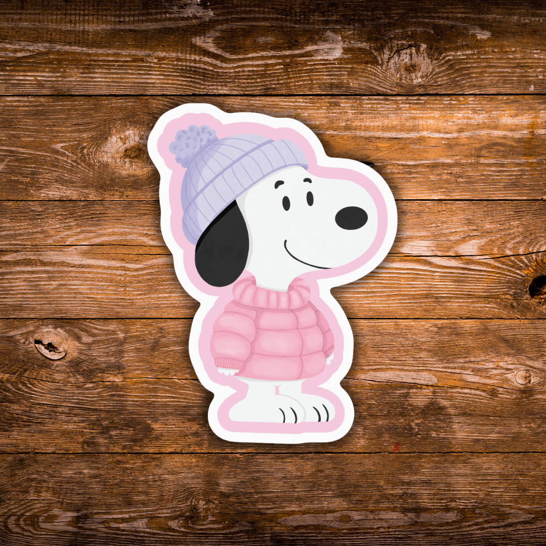 A die-cut sticker of the cartoon dog character wearing a pink sweater and lavender knit hat, placed on a rustic brown wooden surface.