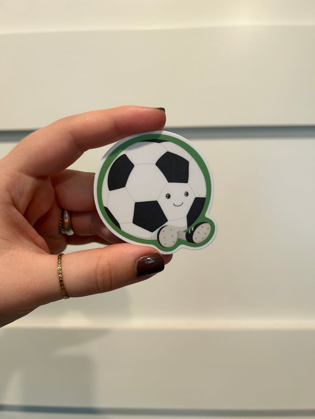 A hand holding a die-cut sticker shaped like a soccer ball with a cute cartoon face, set against a soft-focus background of vertical window panes.
