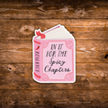 A die-cut sticker shaped like an open romance book, featuring the text 'IN IT FOR THE Spicy Chapters' on a pink cover with a red ribbon and chili pepper accents, displayed on a rustic brown wooden background.