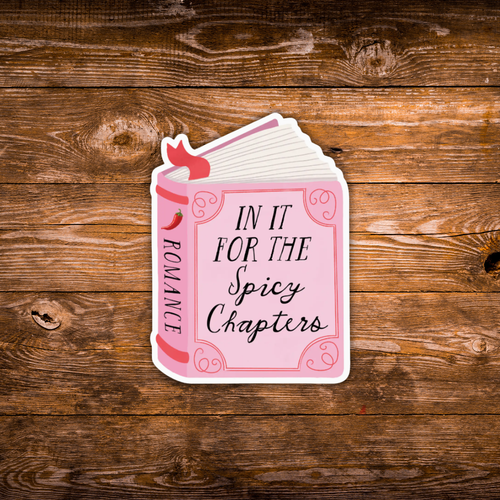 A die-cut sticker shaped like an open romance book, featuring the text 'IN IT FOR THE Spicy Chapters' on a pink cover with a red ribbon and chili pepper accents, displayed on a rustic brown wooden background.