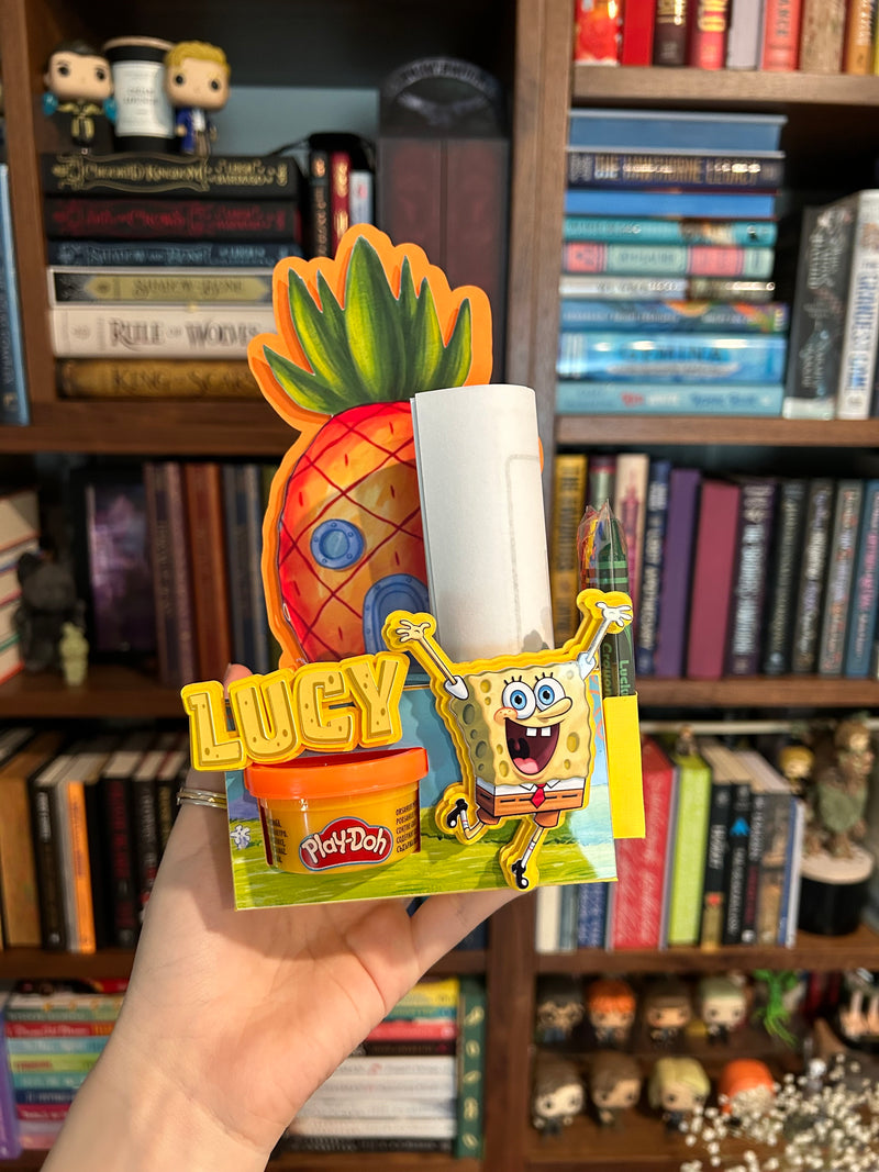 A hand holds a colorful SpongeBob-themed activity box featuring a pineapple house, SpongeBob character, the name 'LUCY', and a Play-Doh container, in front of a bookshelf background.