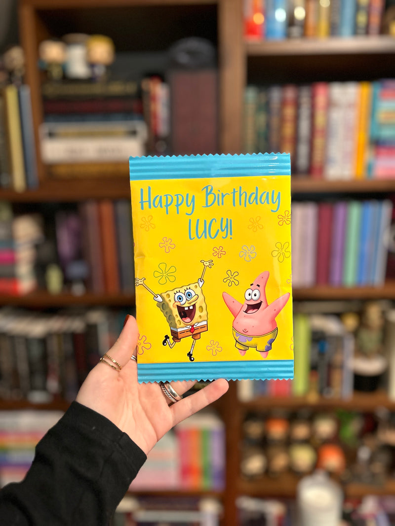 A hand holding a custom SpongeBob-themed birthday chip bag in front of a blurred bookshelf background. The bag is bright yellow with blue borders, featuring SpongeBob and Patrick with the text 'Happy Birthday LUCY!'.