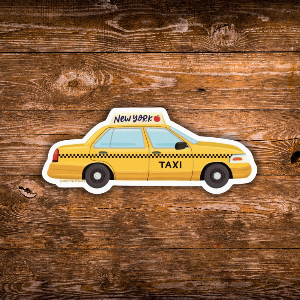 A die-cut vinyl sticker of a yellow New York City taxi cab with black checkered stripes, shown on a rustic brown wooden plank background.