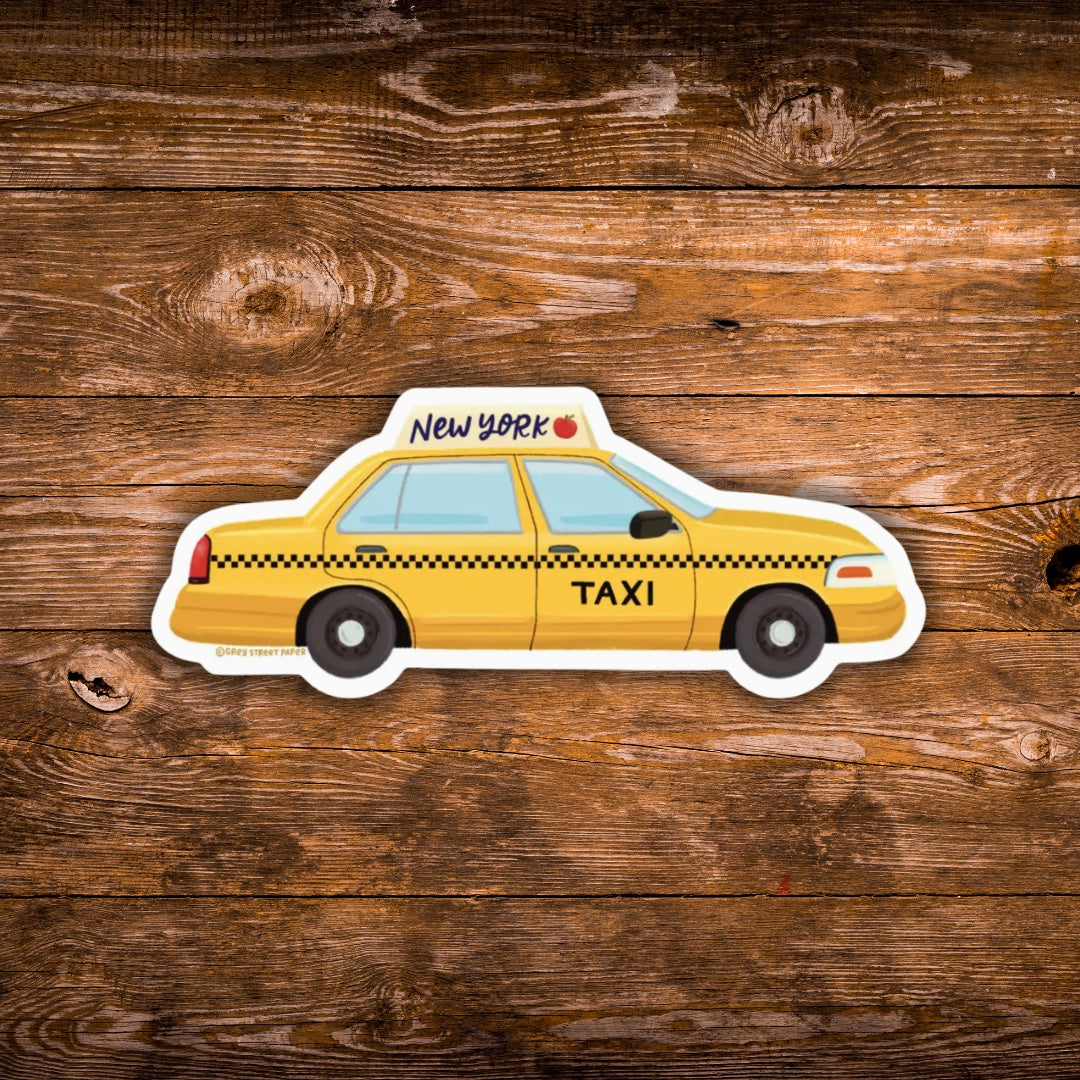 A die-cut vinyl sticker of a yellow New York City taxi cab with black checkered stripes, shown on a rustic brown wooden plank background.