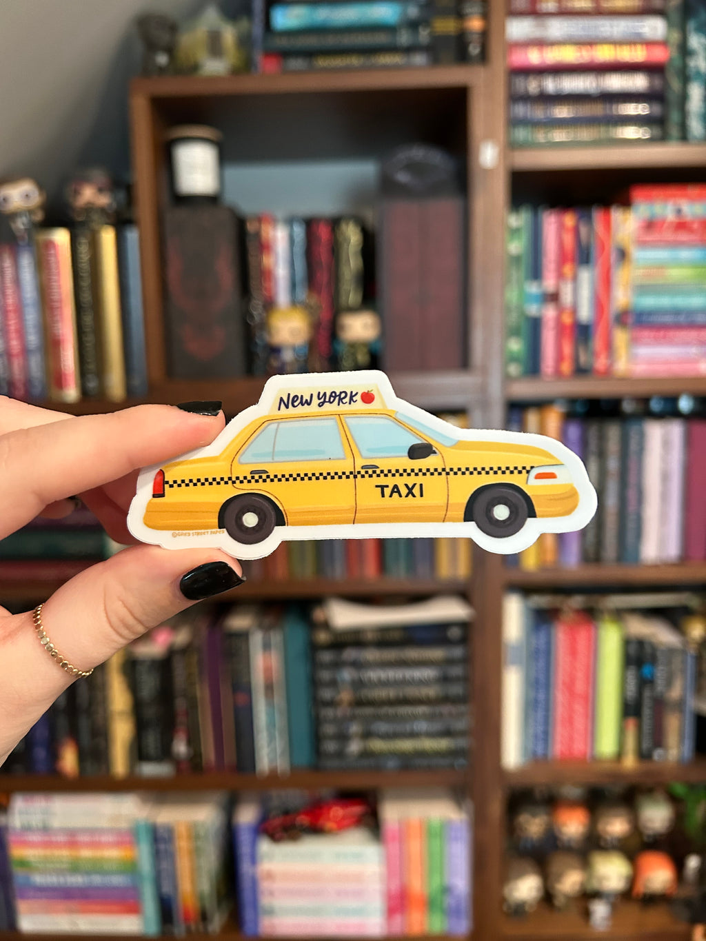 A hand with black nail polish holds a die-cut sticker shaped like a yellow New York City taxi in front of a blurred bookshelf background.
