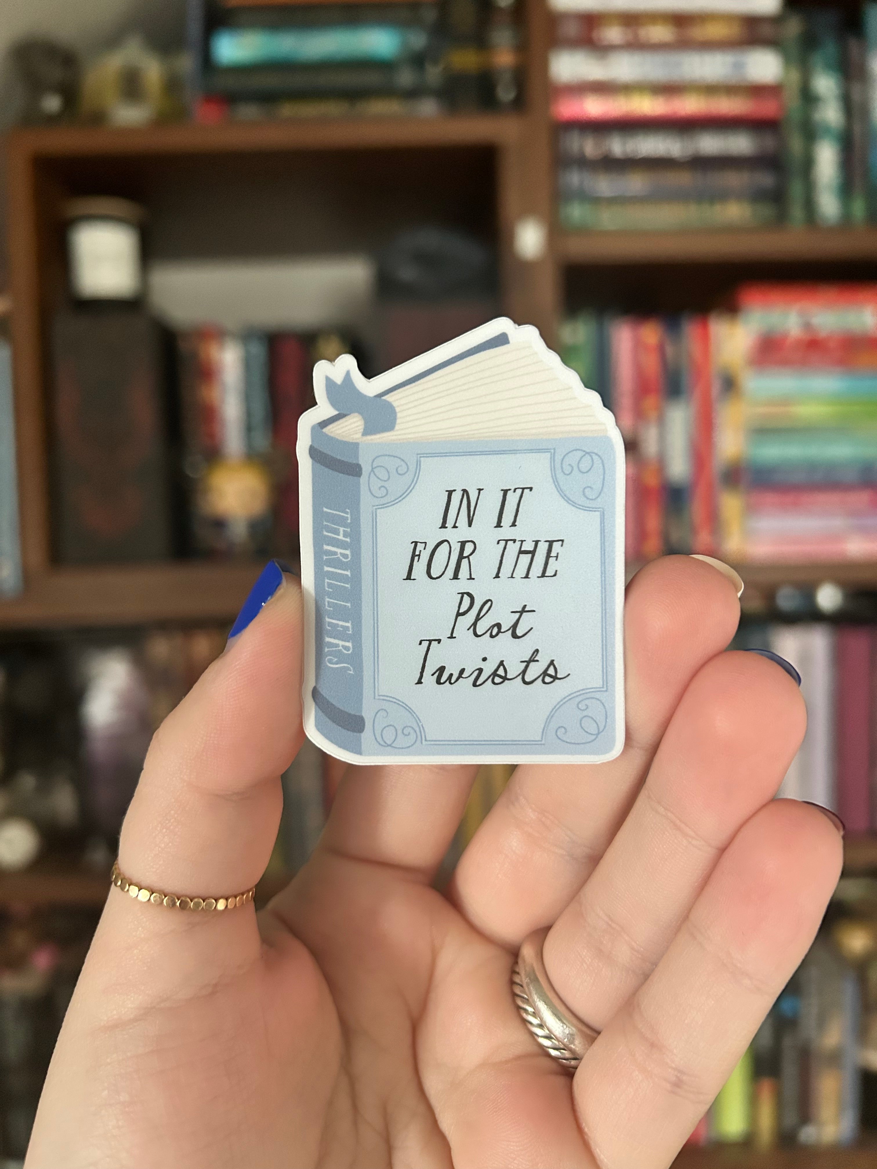 A hand holding a small fanned-out card deck with a single card reading 'IN IT FOR THE Plot Twists' and 'THRILLERS' on the front, set against a blurred bookshelf background.