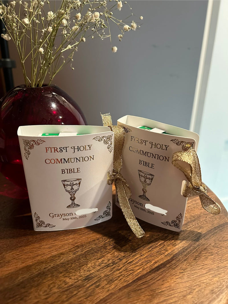 Two personalized First Holy Communion Bible favor boxes tied with gold ribbons, displayed on a wooden table next to a red glass vase with dried baby's breath flowers.