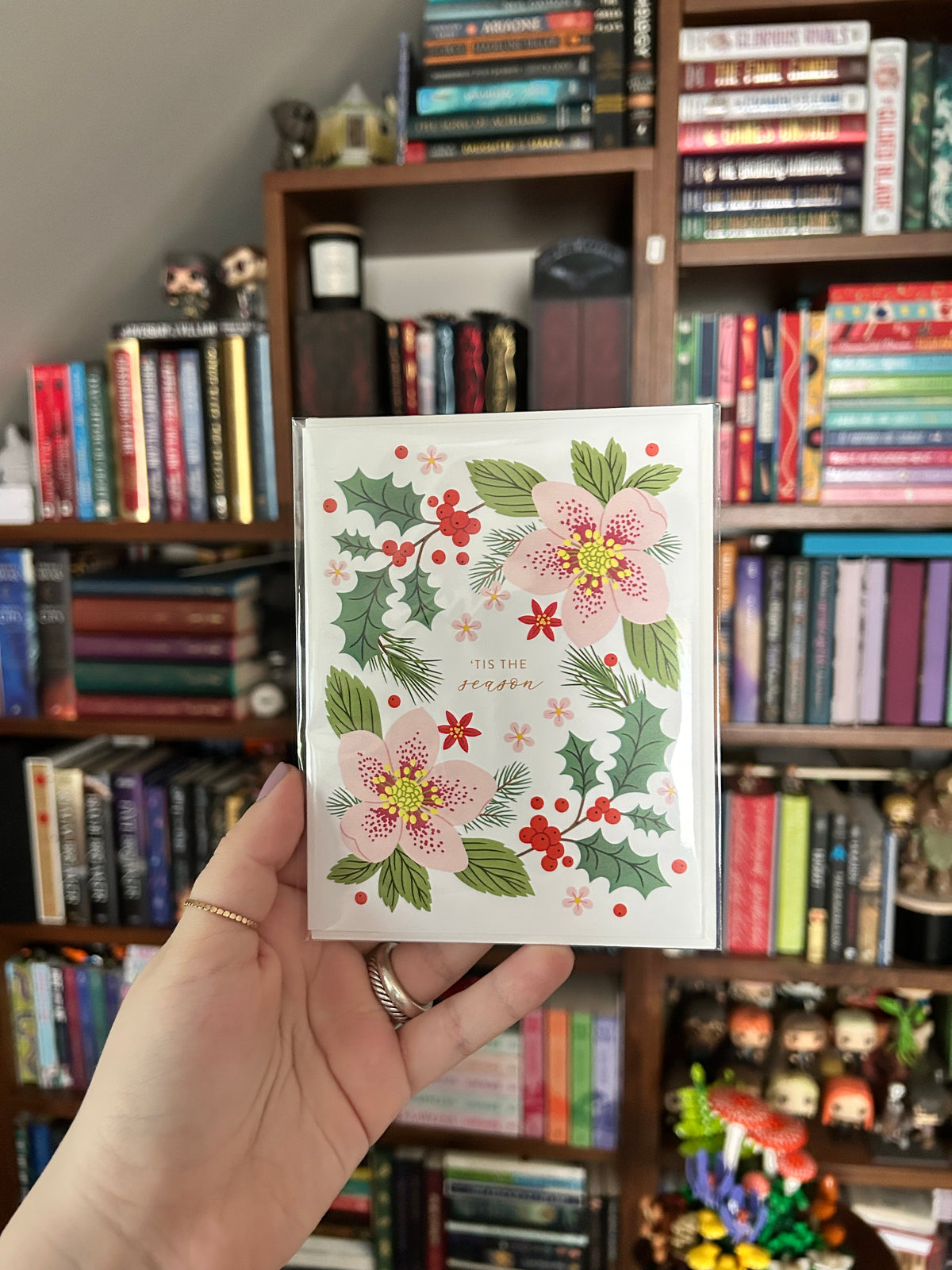 A hand holds a white card featuring a festive illustration of pink flowers, green holly leaves, and red berries, with the text 'TIS THE SEASON' printed on it. The background shows a bookshelf filled with books.