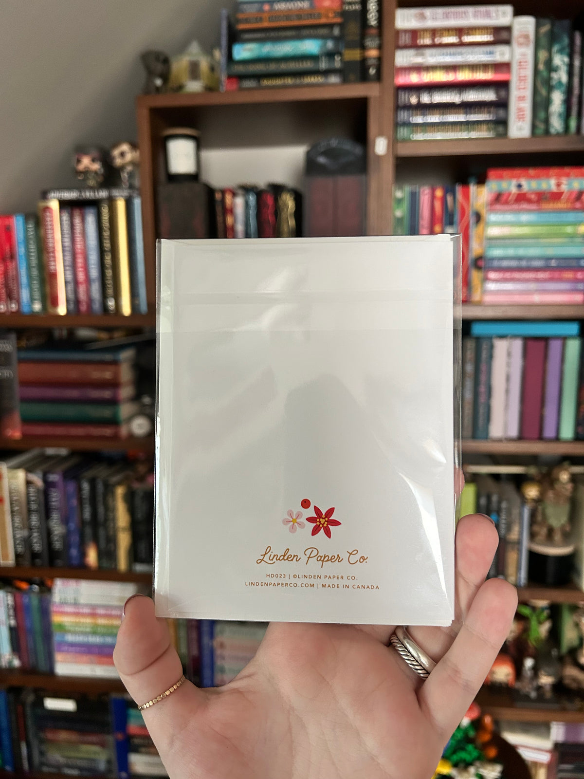 A hand holding a clear plastic-wrapped white card with a small red holly and the text 'Linden Paper Co' in front of a bookshelf filled with books.
