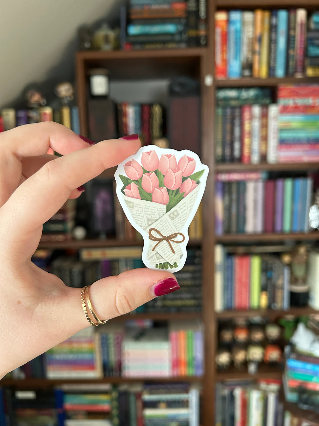 A hand with dark red nail polish holds a small die-cut sticker shaped like a bouquet of pink tulips wrapped in newspaper, with a brown bow, against a blurred background of bookshelves.