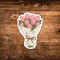 A die-cut sticker depicting a bouquet of pink tulips wrapped in newspaper and tied with a brown bow, shown on a rustic brown wooden background.