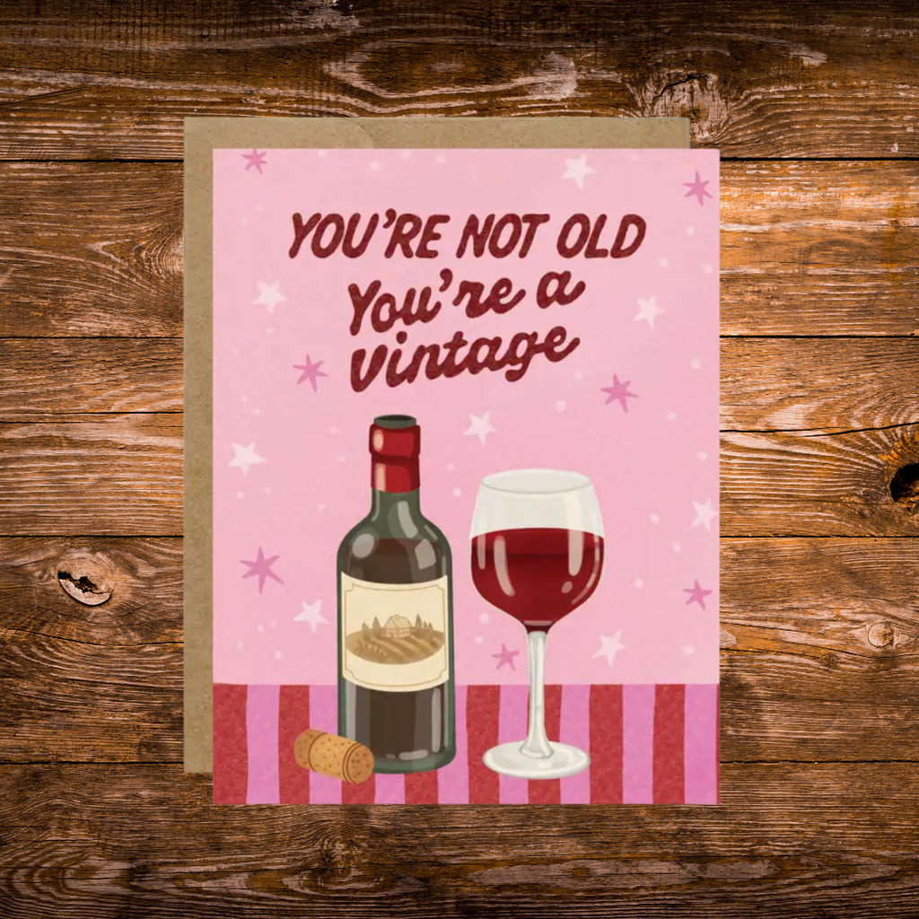Funny birthday greeting card featuring a bottle of red wine and a wine glass with the phrase "You're Not Old, You're a Vintage" on a pink background.