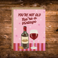 Funny birthday greeting card featuring a bottle of red wine and a wine glass with the phrase "You're Not Old, You're a Vintage" on a pink background.