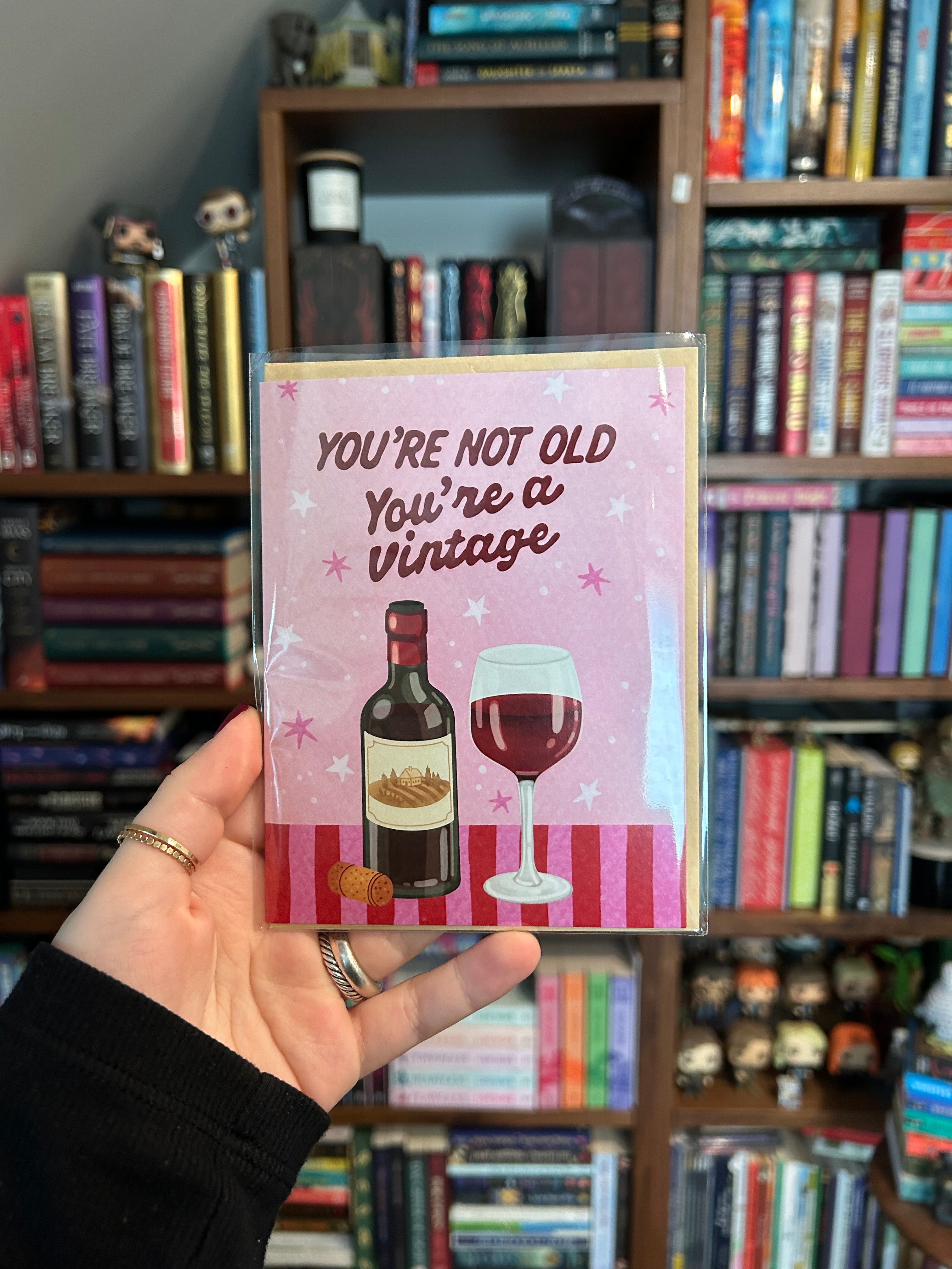 Funny birthday greeting card featuring a bottle of red wine and a wine glass with the phrase "You're Not Old, You're a Vintage" on a pink background.