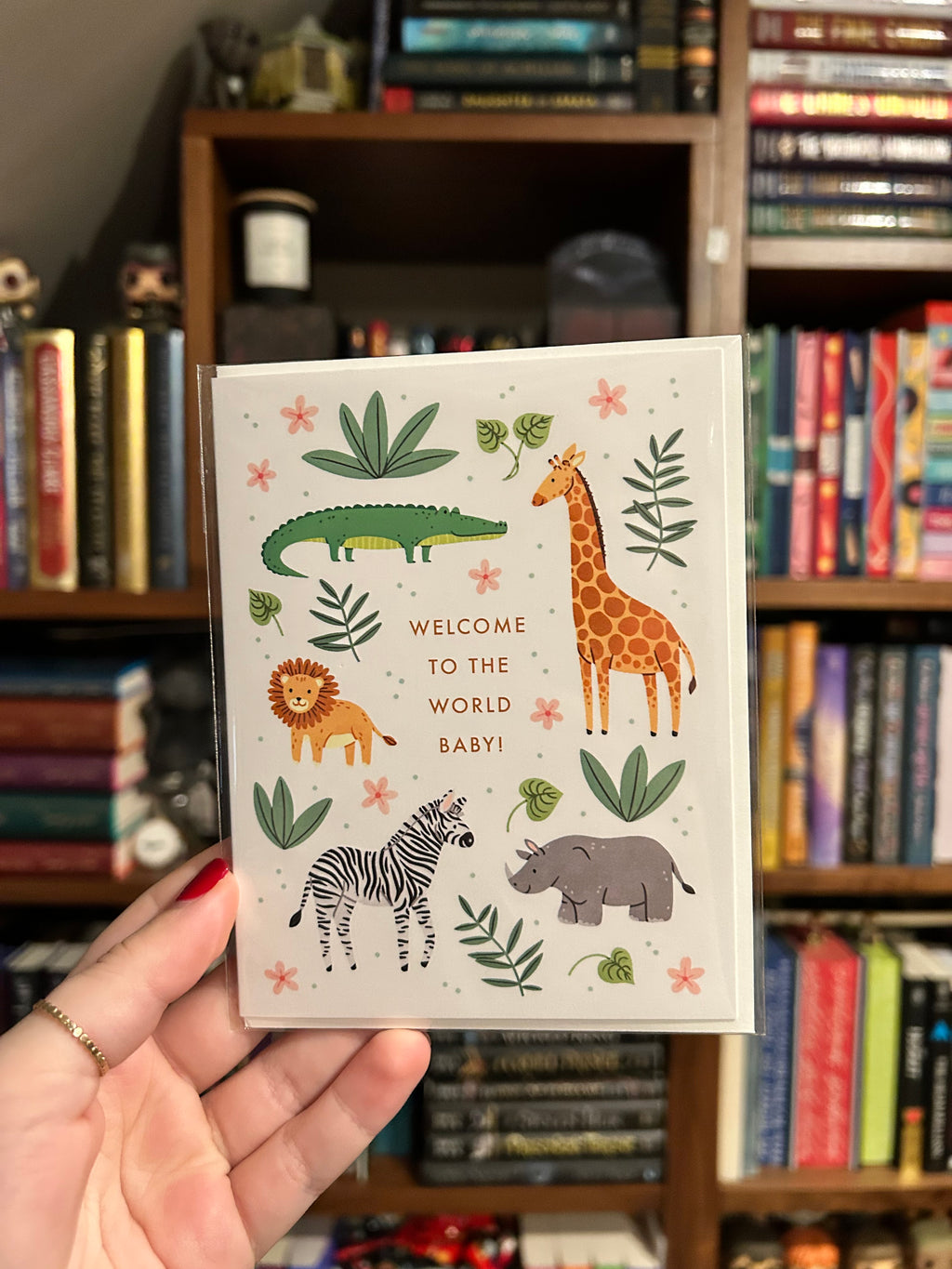 Gender-neutral baby greeting card featuring adorable safari animals including a giraffe, lion, zebra, crocodile, and rhino with greenery and the words "Welcome to the World Baby!"