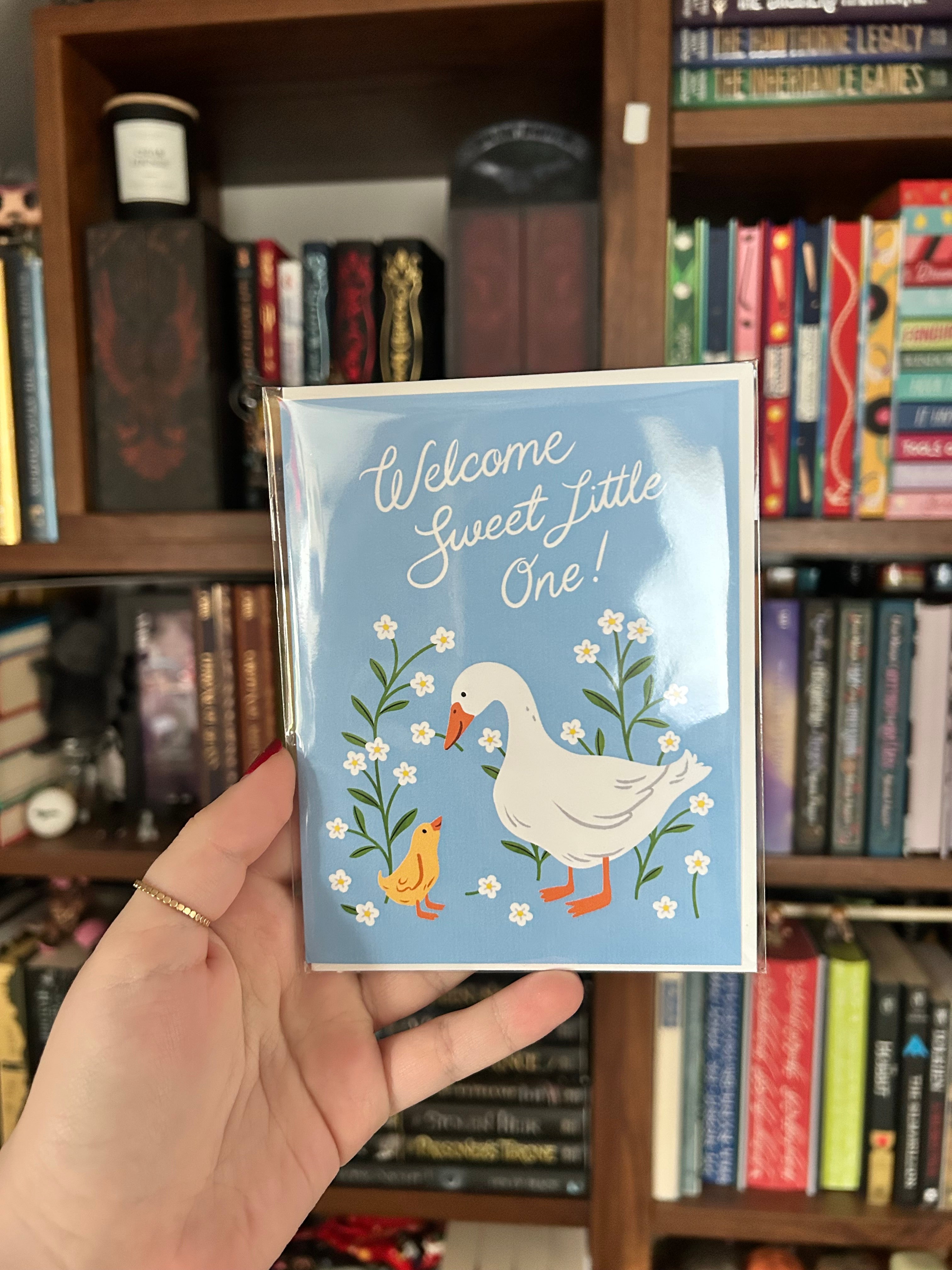Gender-neutral baby greeting card featuring a white goose and yellow gosling surrounded by white wildflowers on a soft blue background with the message "Welcome Sweet Little One!"