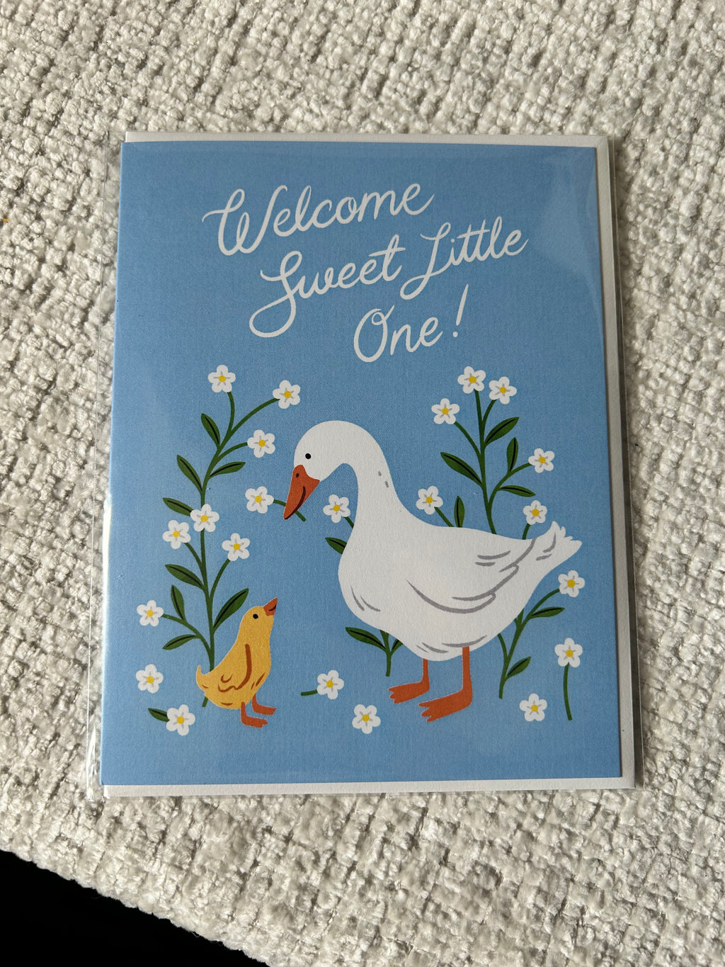 Gender-neutral baby greeting card featuring a white goose and yellow gosling surrounded by white wildflowers on a soft blue background with the message "Welcome Sweet Little One!"