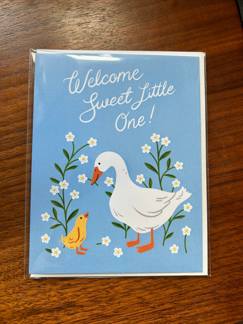 Gender-neutral baby greeting card featuring a white goose and yellow gosling surrounded by white wildflowers on a soft blue background with the message "Welcome Sweet Little One!"