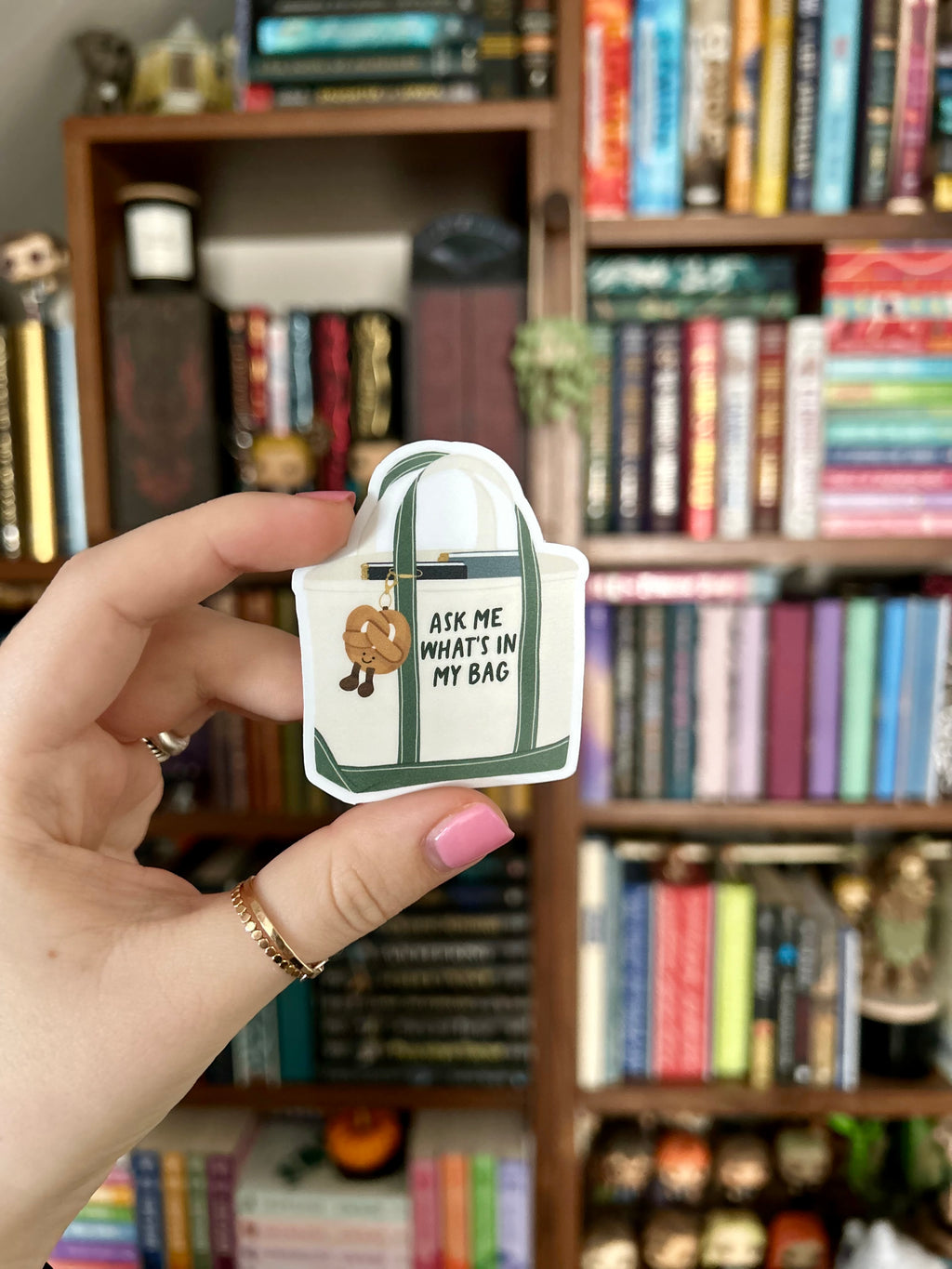 A hand with pink nail polish holds a small enamel pin shaped like a white tote bag with green handles and straps. The pin features a small brown cartoon character and the text 'ASK ME WHAT'S IN MY BAG'.