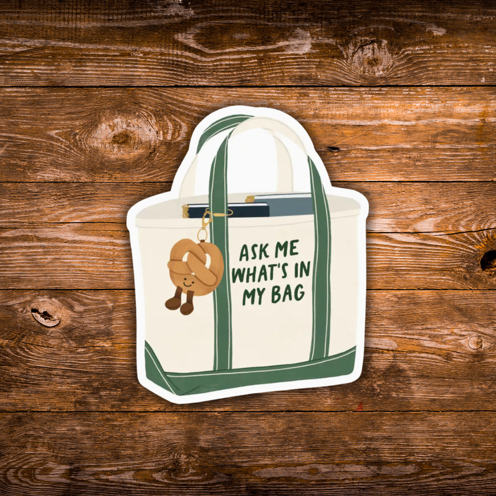 A die-cut sticker of a cream canvas tote bag with green handles and trim, featuring the text 'ASK ME WHAT'S IN MY BAG' and a pretzel charm, displayed on a rustic brown wood background.