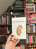 White Christmas greeting card featuring a smiling green pickle wearing a Santa hat and red-and-white scarf with the phrase “Have a Very Merry Dill-mas.” 
