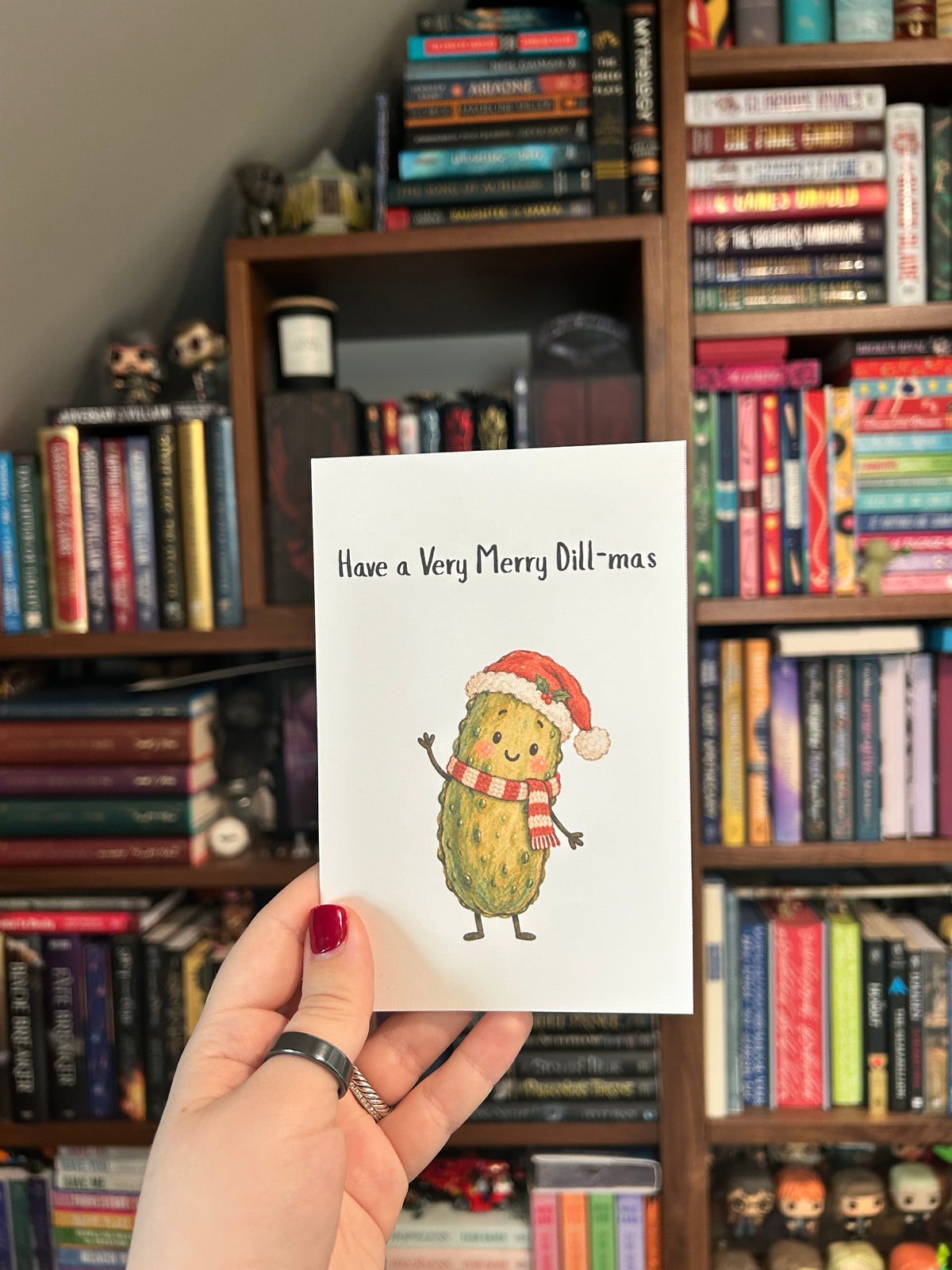 White Christmas greeting card featuring a smiling green pickle wearing a Santa hat and red-and-white scarf with the phrase “Have a Very Merry Dill-mas.” 
