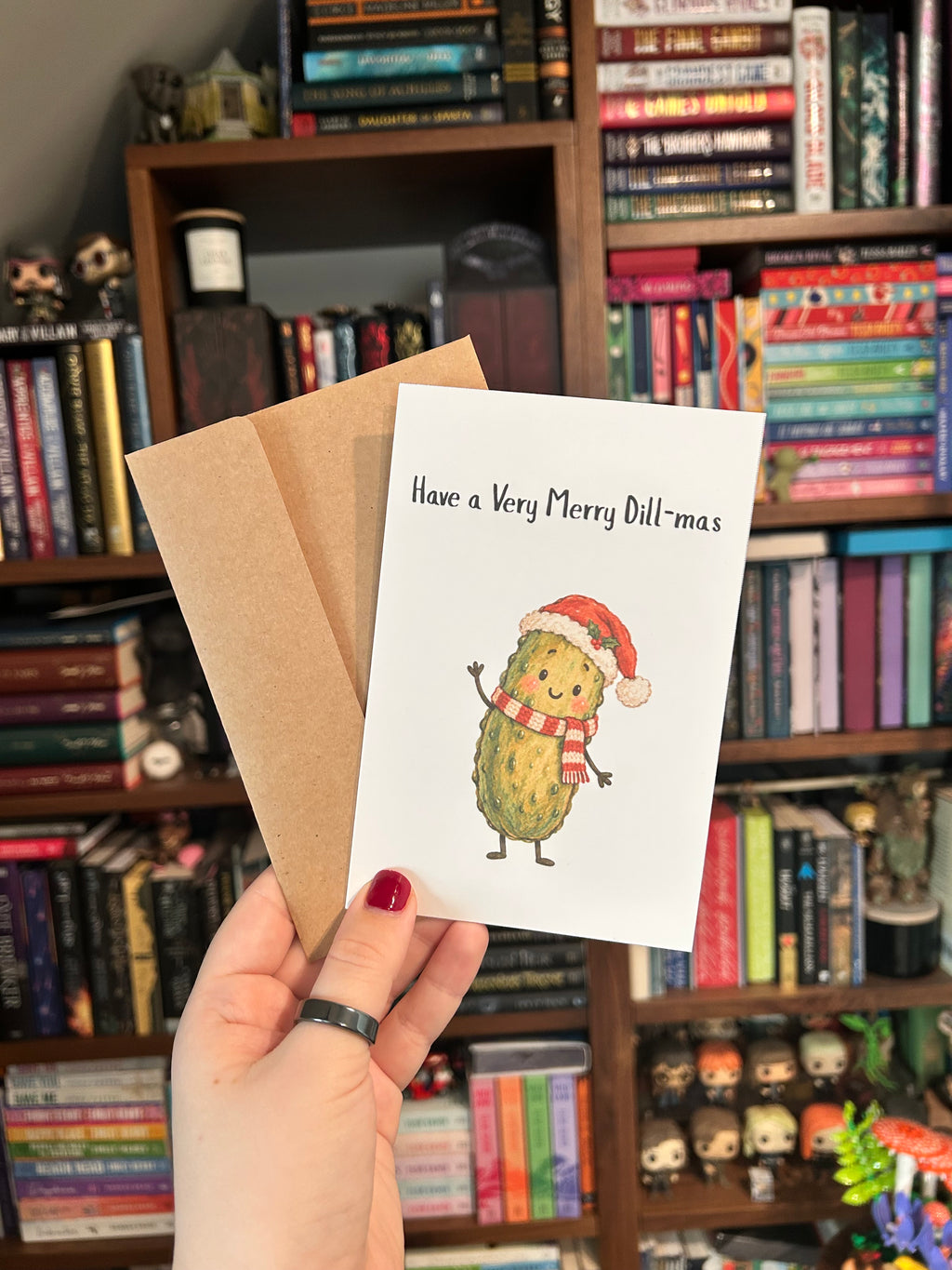 White Christmas greeting card featuring a smiling green pickle wearing a Santa hat and red-and-white scarf with the phrase “Have a Very Merry Dill-mas” with a kraft envelope.