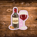 A die-cut sticker featuring an illustrated wine bottle, filled wine glass, and cork on a pink background, shown against a rustic brown wooden plank backdrop.
