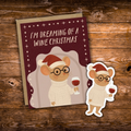 A Christmas-themed mouse sticker and matching greeting card on a rustic wooden background. The card features a cartoon mouse wearing glasses, a white knit sweater, and a red Santa hat, holding a glass of red wine, with the text 'I'M DREAMING OF A WINE CHRISTMAS'.