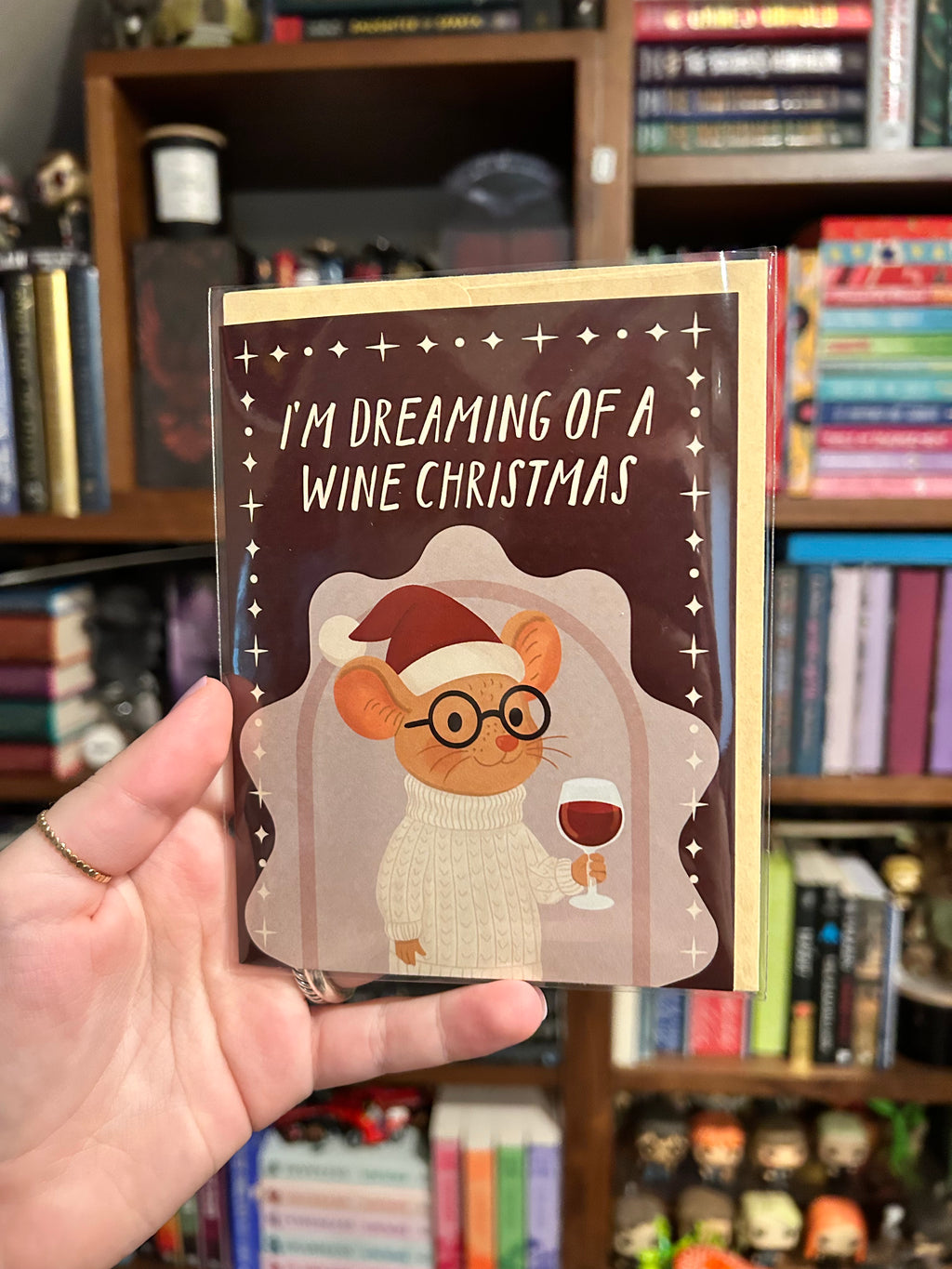 A hand holds a Christmas greeting card in front of a bookshelf. The card has a dark brown background with the text 'I'M DREAMING OF A WINE CHRISTMAS' and an illustration of a mouse wearing a Santa hat and glasses while holding a glass of red wine.
