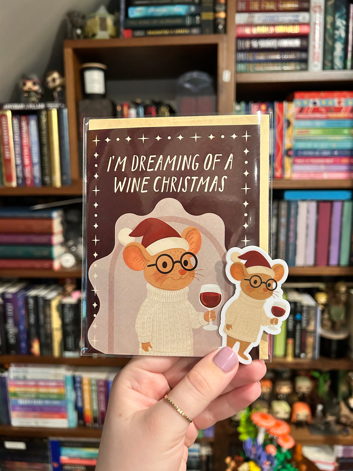 A hand holds a Christmas-themed greeting card and matching sticker featuring cartoon mice wearing Santa hats and glasses, with the text 'I'M DREAMING OF A WINE CHRISTMAS', against a blurred background of bookshelves.