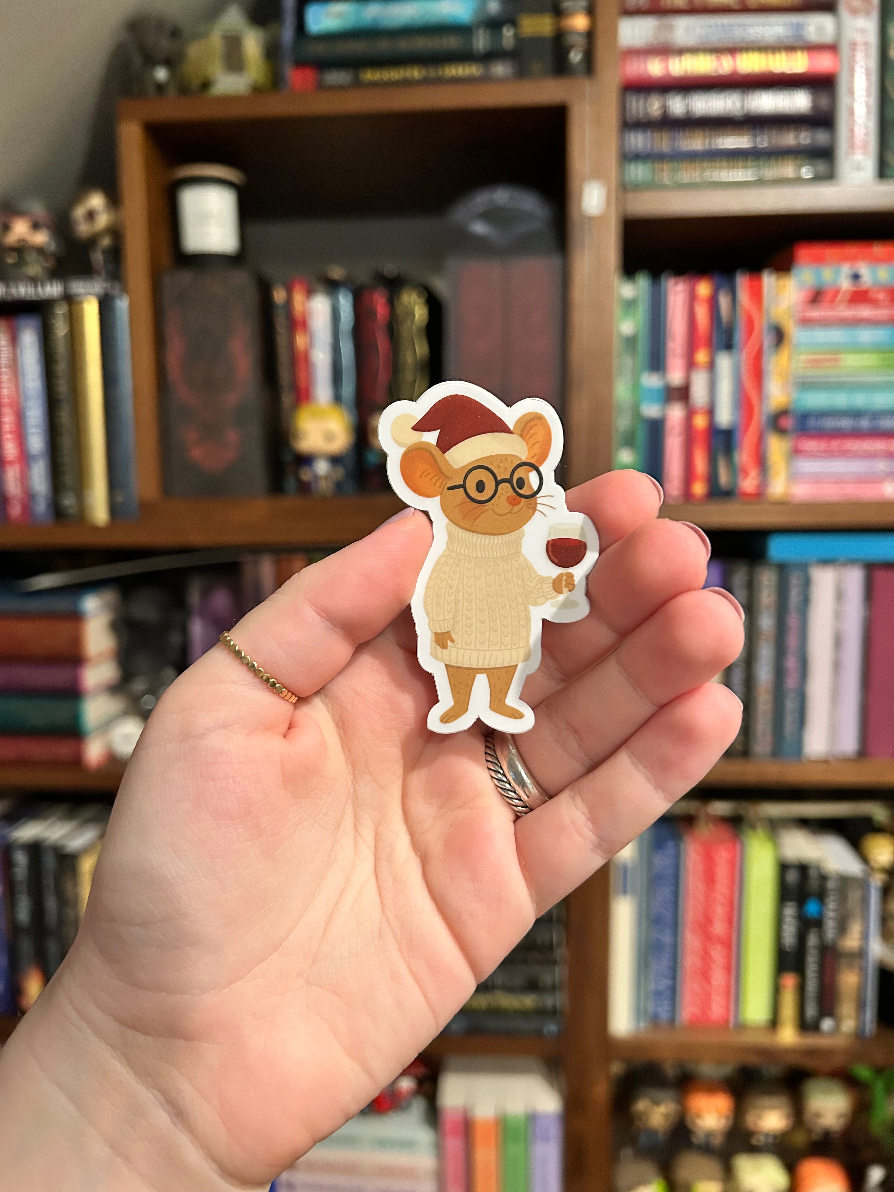 A hand holding a die-cut sticker of a cute cartoon mouse wearing a Santa hat and glasses, with a blurred bookshelf in the background.