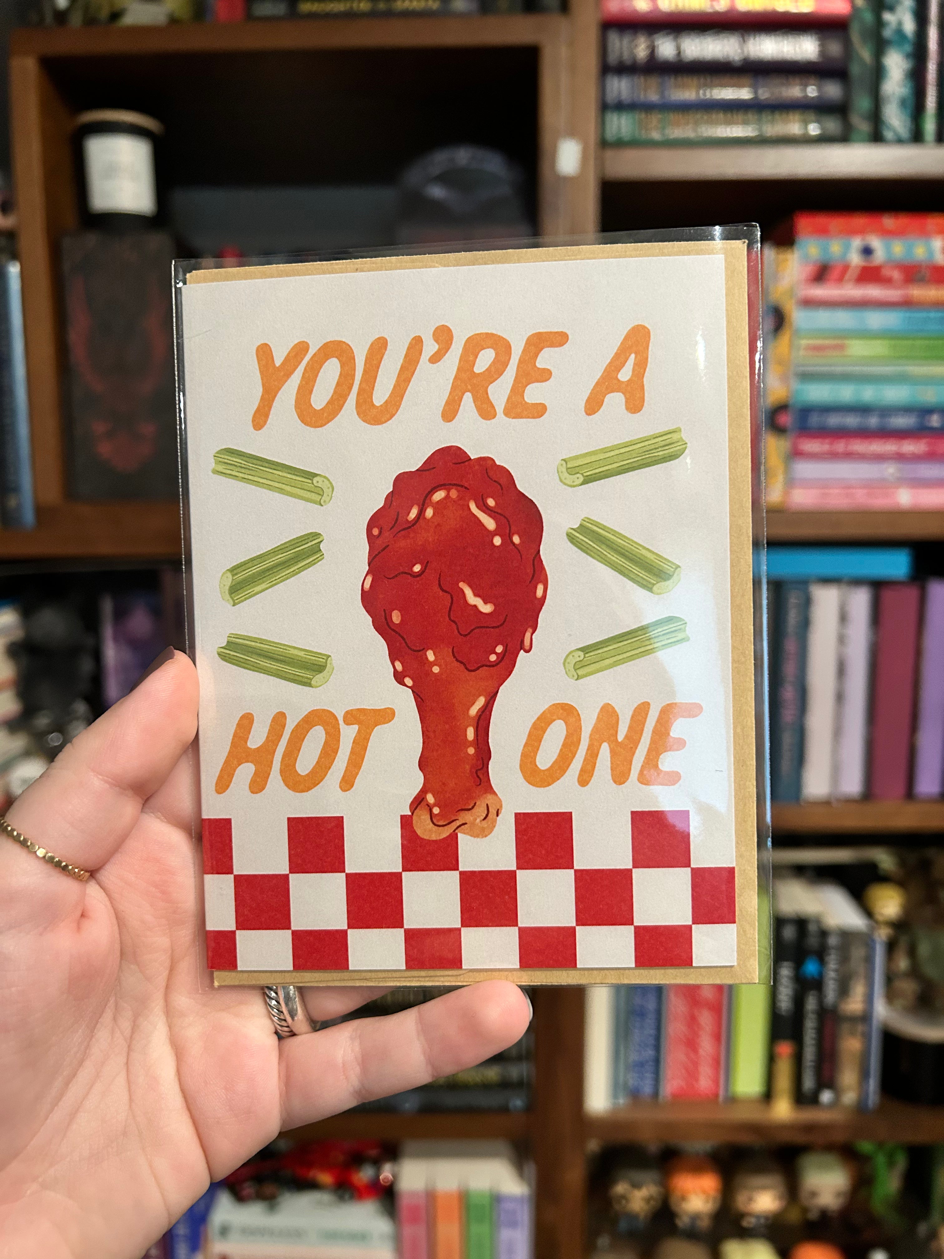A hand holds a square greeting card in a clear sleeve in front of a bookshelf. The card features a chicken leg drumstick covered in red sauce, celery sticks, and the text 'YOU'RE A HOT ONE' in orange.