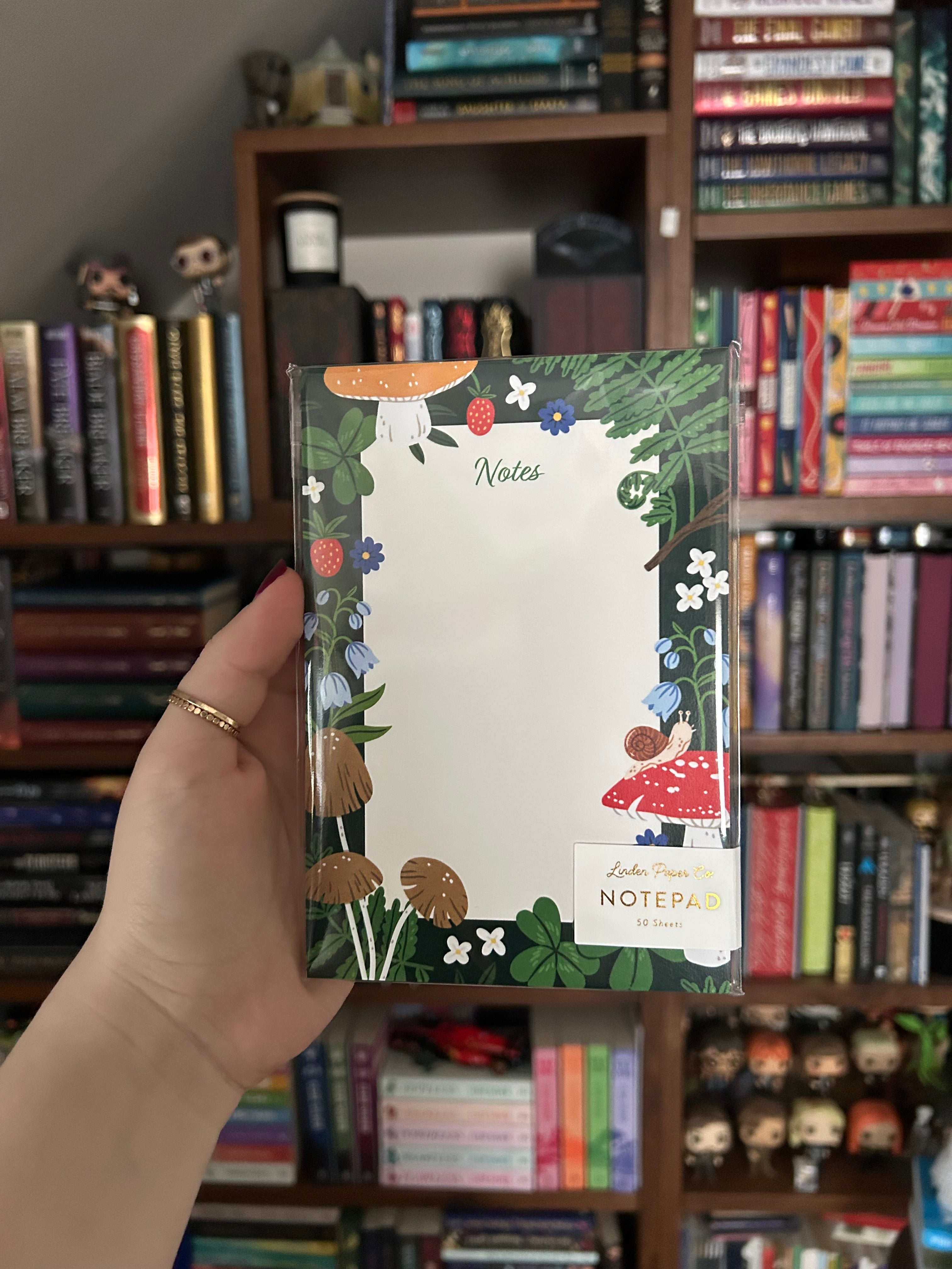 Woodland-themed notepad featuring mushrooms, ferns, strawberries, clovers, wildflowers, and a small snail surrounding a large blank writing area on a dark green background.