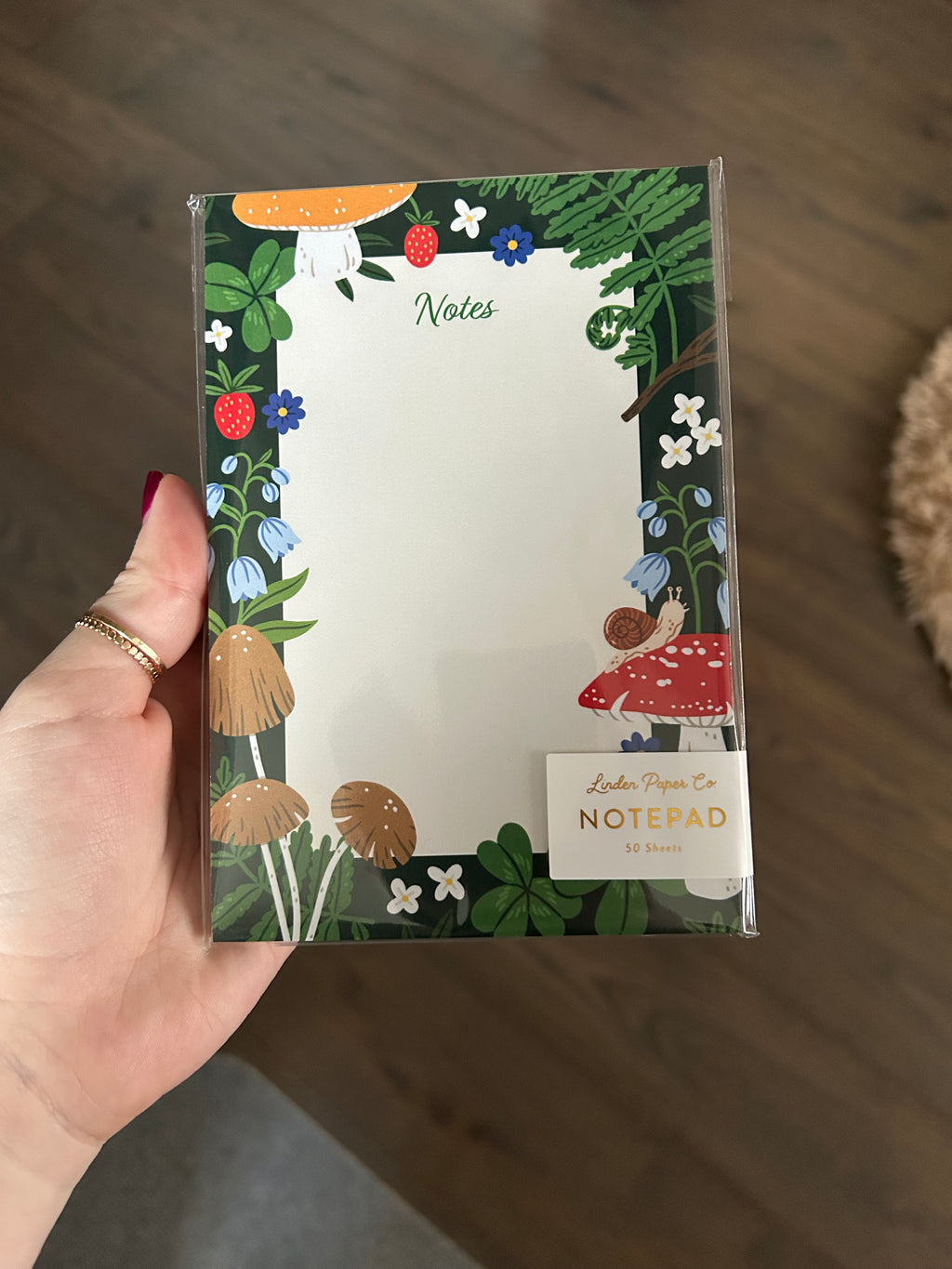 Woodland-themed notepad featuring mushrooms, ferns, strawberries, clovers, wildflowers, and a small snail surrounding a large blank writing area on a dark green background.