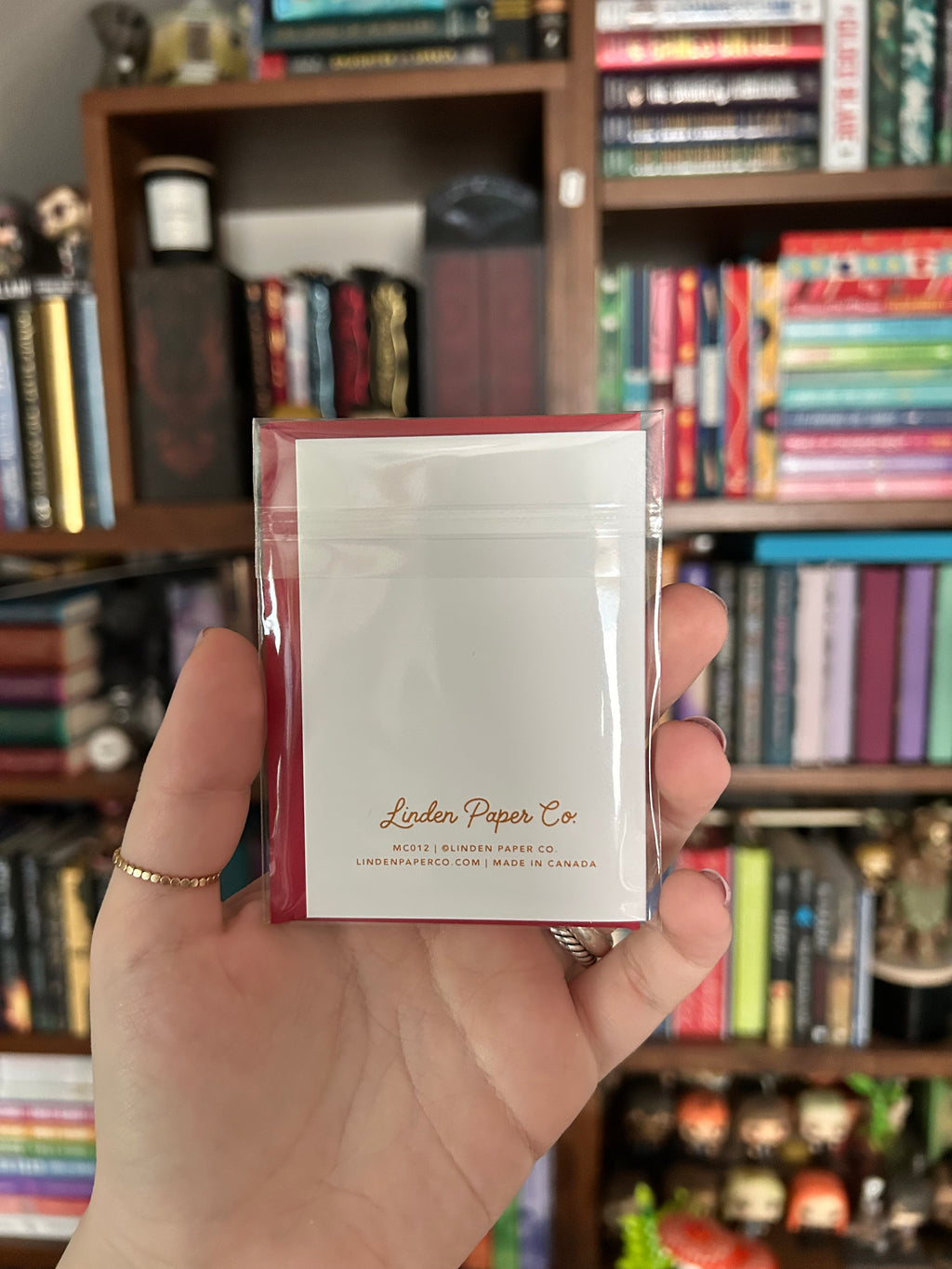 A hand holding up the back of a Christmas card with the brand name 'Linden Paper Co' printed on it, set against a blurred background of bookshelves.