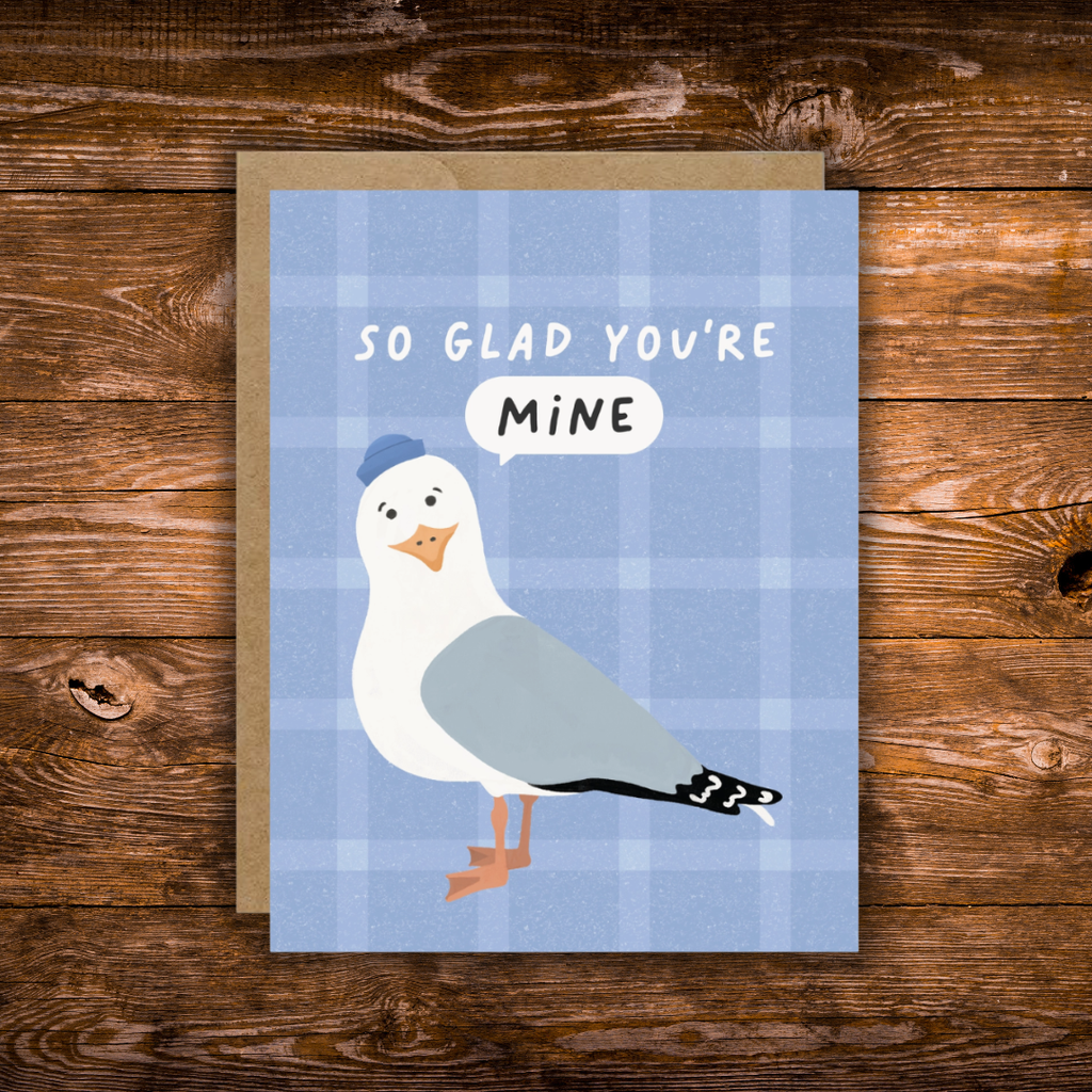 A square greeting card with a blue plaid background, featuring an illustrated seagull wearing a blue hat and a speech bubble that says 'MINE', with the card text 'SO GLAD YOU'RE MINE'. The card is placed on top of a kraft envelope on a rustic wooden surface.