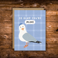 A square greeting card with a blue plaid background, featuring an illustrated seagull wearing a blue hat and a speech bubble that says 'MINE', with the card text 'SO GLAD YOU'RE MINE'. The card is placed on top of a kraft envelope on a rustic wooden surface.
