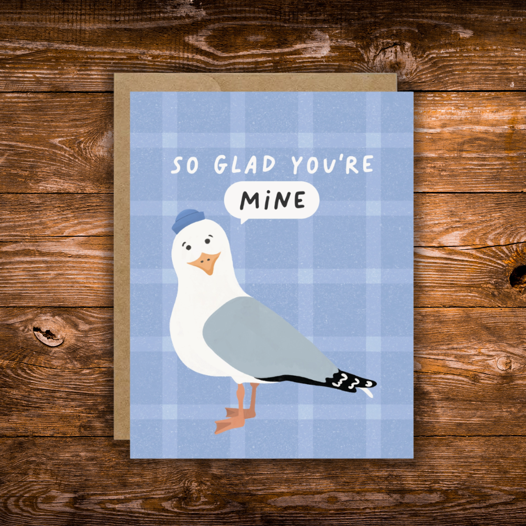 A square greeting card with a blue plaid background, featuring an illustrated seagull wearing a blue hat and a speech bubble that says 'MINE', with the card text 'SO GLAD YOU'RE MINE'. The card is placed on top of a kraft envelope on a rustic wooden surface.