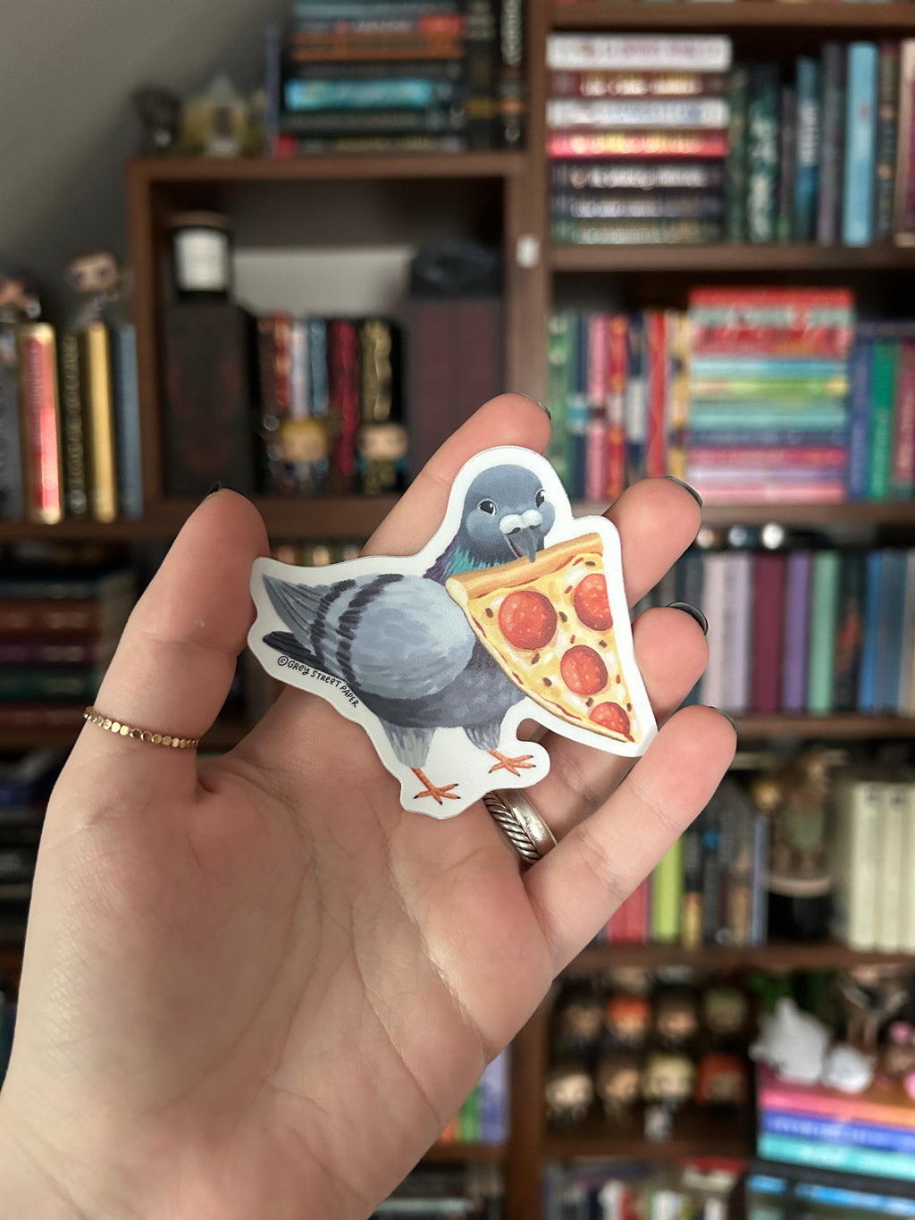 A hand holding a die-cut sticker featuring a gray pigeon eating a slice of pepperoni pizza, with a blurred bookshelf in the background.