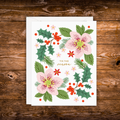 A square white greeting card with a festive floral illustration, placed on top of a white envelope over a rustic brown wooden surface. The card features pink blossoms, green holly leaves, red berries, pine branches, and the words “TIS THE season” in gold script.