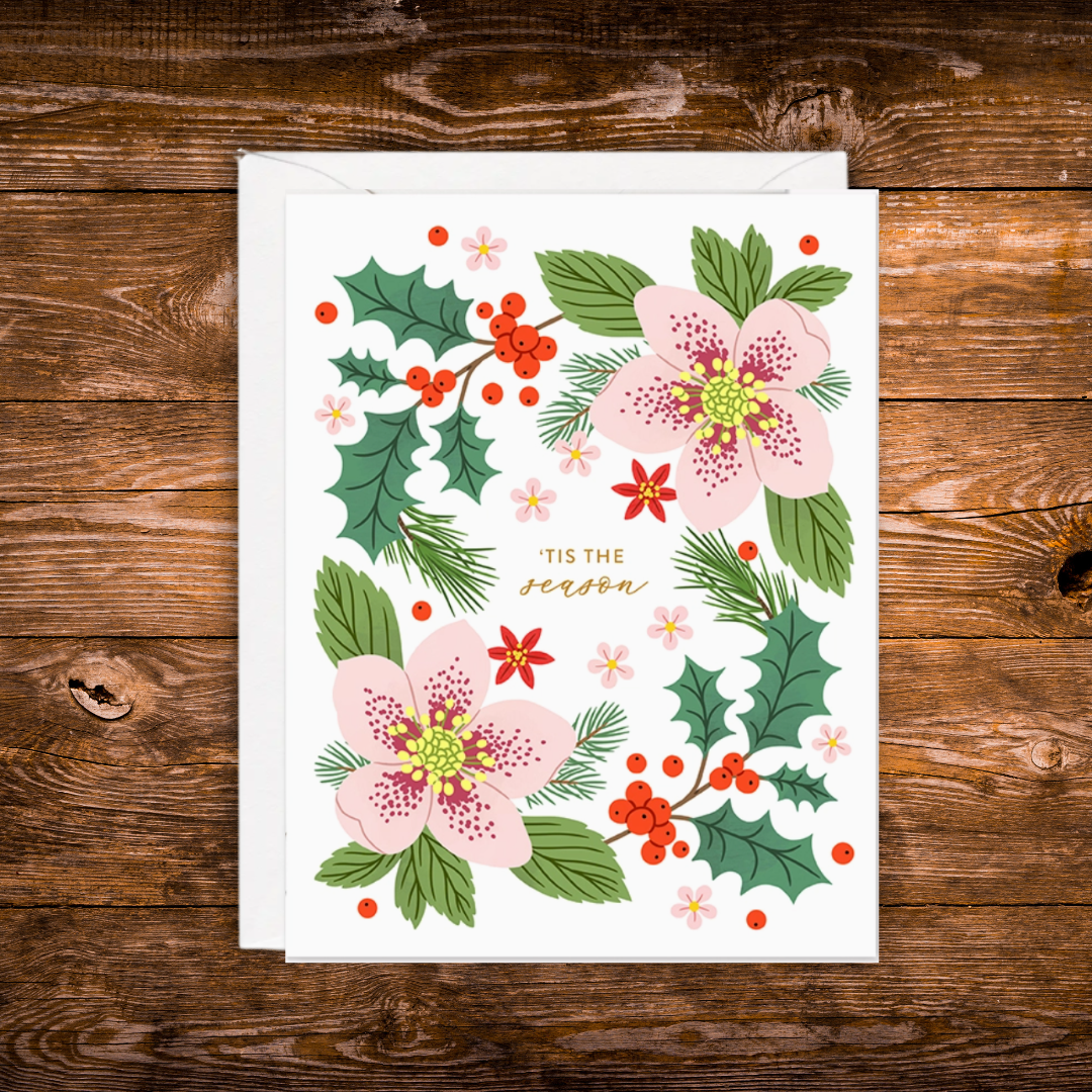 A square white greeting card with a festive floral illustration, placed on top of a white envelope over a rustic brown wooden surface. The card features pink blossoms, green holly leaves, red berries, pine branches, and the words “TIS THE season” in gold script.